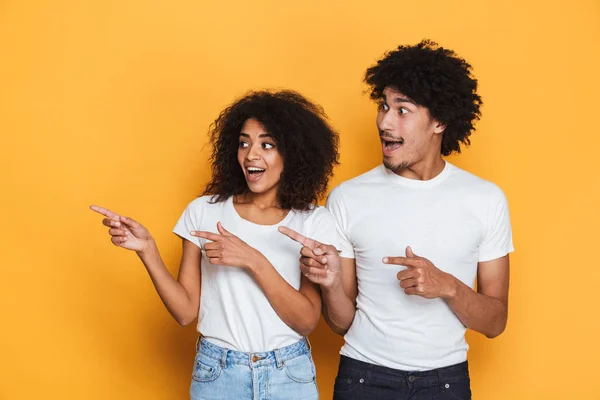 Portrait of a happy young afro american couple pointing fingers away at copy space isolated over yellow background