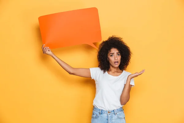 Portrait of a confused young african woman holding empty speech bubble isolated over yellow background