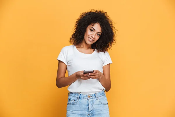 黄色の背景に分離された携帯電話を保持している笑顔の若いアフリカ女性の肖像画 — ストック写真