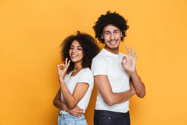 Portrait of a smiling young afro american couple standing and showing ok gesture isolated over yellow background