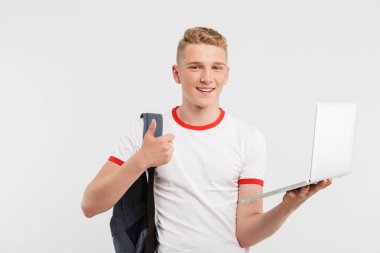 Neşeli erkek öğrenci tişört ve sırt çantası açık laptop beyaz arka plan üzerinde izole tutarken başparmak gösterilmesini görüntüsünü