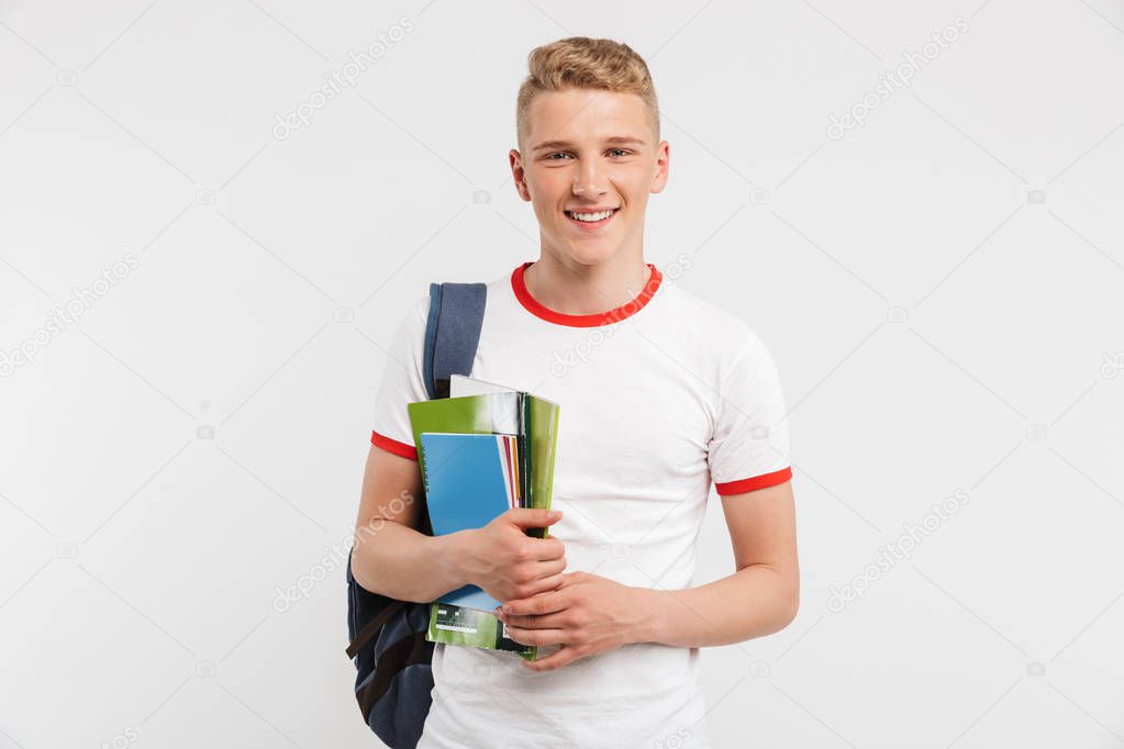 Image of european student boy wearing backpack smiling and posing at camera with colorful exercise books in hands isolated over white background