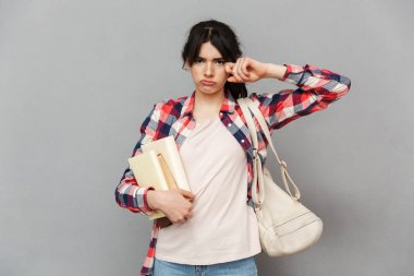 Kitap tutan gri arka plan duvardan izole duran genç bayan öğrenci üzücü görüntüsünü sıktı.