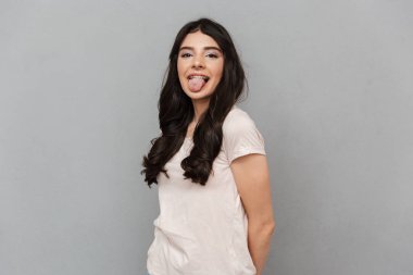 Sevimli güzel genç bayan dil gösterilen gri arka plan duvardan izole ayakta görüntüsünü.