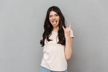 Heyecanlı güzel genç bayan gri arka plan duvardan izole ayakta görüntüsü. Kamera gösteren rock jest arıyorsunuz.