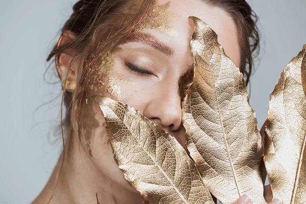 Harika esmer kadının yumuşak sağlıklı deri ve altın makyaj ile görüntü güzellik altın yaprak tutan gri arka plan üzerinde izole. Gözleri kapalı.