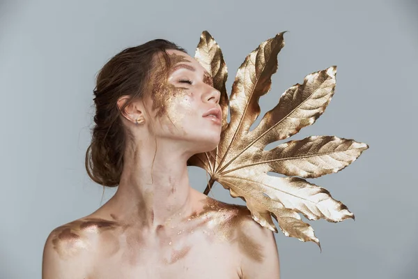 Harika esmer kadının yumuşak sağlıklı deri ve altın makyaj ile görüntü güzellik altın yaprak tutan gri arka plan üzerinde izole. Gözleri kapalı.