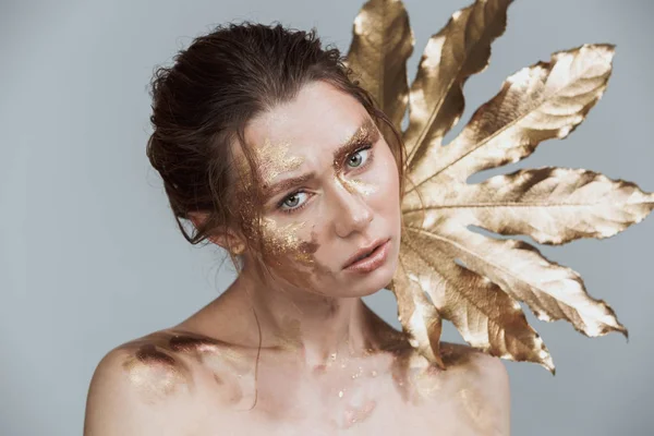 Altın yaprak tutan kamera seyir gri arka plan üzerinde şaşırtıcı esmer kadının yumuşak sağlıklı deri ve altın makyaj ile görüntü güzellik izole.