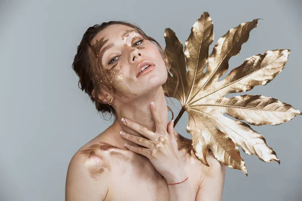Altın yaprak tutan kamera seyir gri arka plan üzerinde şaşırtıcı esmer kadının yumuşak sağlıklı deri ve altın makyaj ile görüntü güzellik izole.