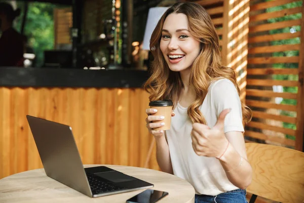 Dizüstü bilgisayar ile oturan ve kahve Kupası'nda bir kafede açık havada tutan jest Yaşasın gösterilen bir gülümseyen kız portresi