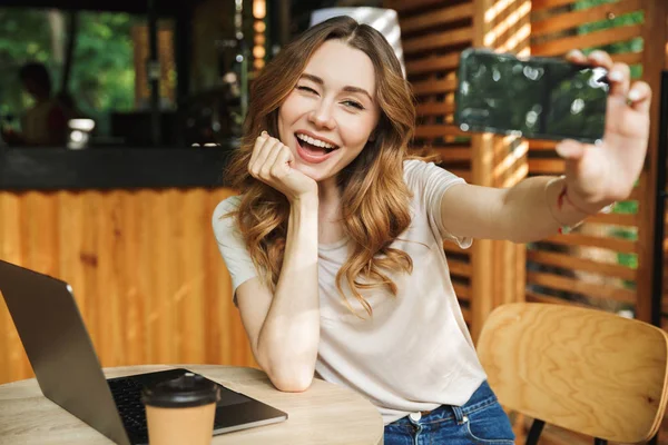 Selfie cep telefonu ile bir kafe açık havada, dizüstü bilgisayar ile otururken alarak oynak bir genç kız portresi