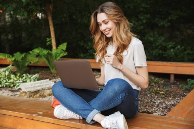 Açık havada bankta otururken dizüstü bilgisayar kullanan genç kız gülümseyerek