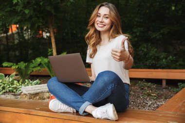 Süre bir tezgah açık havada üzerinde oturan ve başparmak gösterilmesini dizüstü bilgisayar kullanan genç kız gülümseyerek