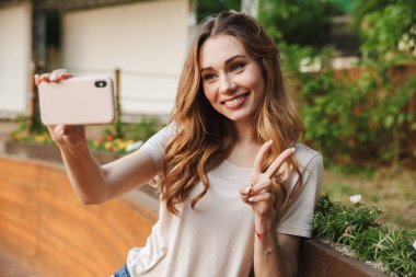 Açık havada ayakta iken selfie alarak rahat kıyafetler içindeki gülümseyen genç kız