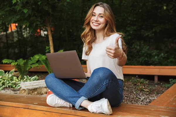 Süre bir tezgah açık havada üzerinde oturan ve başparmak gösterilmesini dizüstü bilgisayar kullanan genç kız gülümseyerek