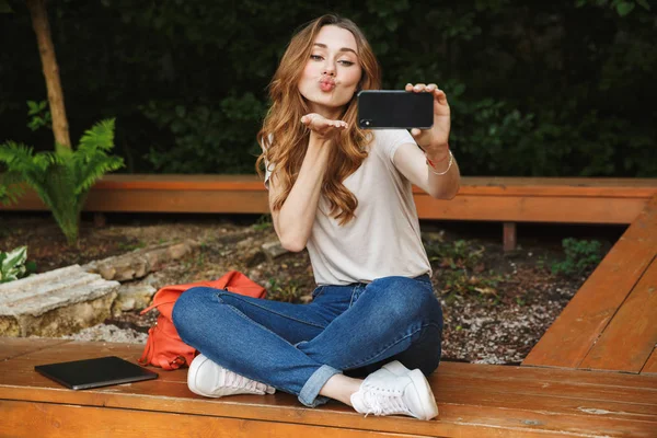 Bir bankta açık havada oturma ve öpücük gönderme selfie alarak güzel genç kız