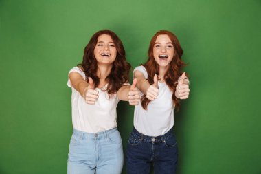 İki neşeli Kızıl saçlı kadınlar 20'li yaşlarda kameraya gülümseyen ve başparmak yeşil arka plan üzerinde izole gösterilmesini rahat kıyafet fotoğrafı