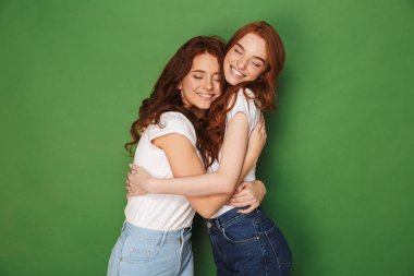 İki çekici zencefil kızlar 20s gülümseyen ve üzerinde yeşil arka plan izole kapalı gözlerle birbirlerine sarılma rahat kıyafet içinde