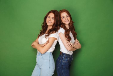 Kameraya gülümseyen ve arka arkaya silah ile ayakta rahat kıyafet Kızıl saçlı iki mutlu kızlar 20s izole yeşil arka plan üzerinde çarpı işareti