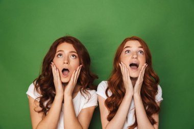 İki Kızıl saçlı kadınlar 20'li yaşlarda beyaz t-shirt yanakları ile açık ağız dokunmaktan ve yukarı doğru seyir üzerinde yeşil arka plan izole fotoğrafı