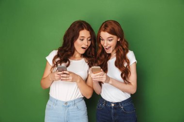 Kızıl saçlı yeşil arka plan üzerinde izole sürpriz ile cep telefonları kullanarak iki sevimli kadın portresi