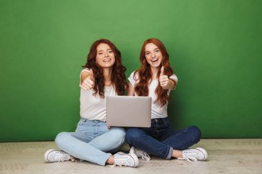 İki dizüstü bilgisayar ile bir katta oturan ve üzerinde yeşil arka plan izole jest Yaşasın gösterilen genç Kızıl saçlı kız gülümseyerek portresi