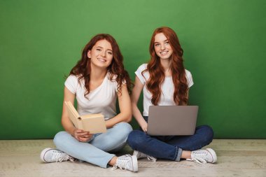 Dizüstü bilgisayar ve kağıt kitap üzerinde yeşil arka plan izole bir katta oturan iki mutlu genç Kızıl saçlı kız portresi