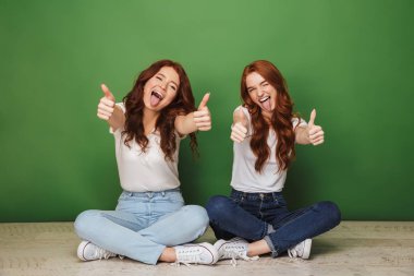 İki genç Kızıl saçlı kız bir katta oturan ve üzerinde yeşil arka plan izole jest Yaşasın gösterilen gülümseyen portresi