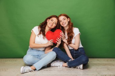 İki çekici Kızıl saçlı kız 20'li yaşlarda kot pantolon ile geçti ve kırmızı kağıt kalp holding bacaklar yere oturma portre üzerinde yeşil arka plan izole