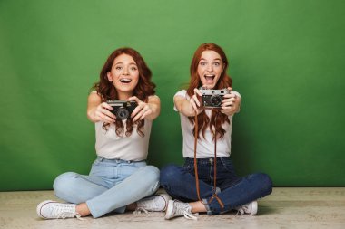 Yeşil arka plan üzerinde iki neşeli Kızıl saçlı kız 20'li yaşlarda geçti ve retro kameralar gösteren bacaklar ile katta oturan Jeans fotoğraf izole