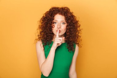 Portrait of a smiling curly redhead woman showing silence gesture isolated over yellow background clipart