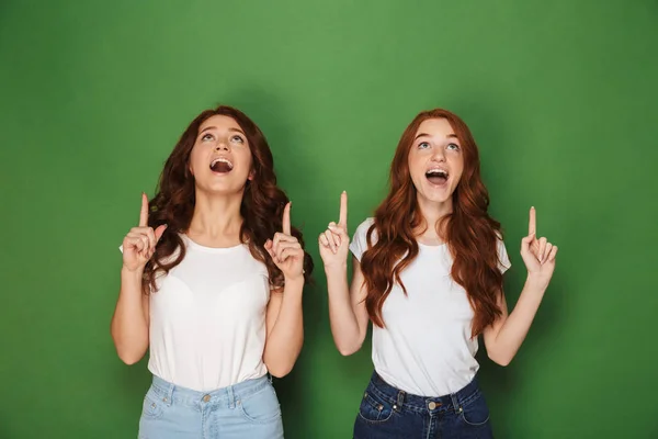 Portrait of two pretty women with red hair in white t-shirts smiling and pointing fingers upward in excitement isolated over green background