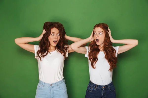 Portrait of two scared or uptight women with red hair in white t-shirts looking at each other and covering ears isolated over green background