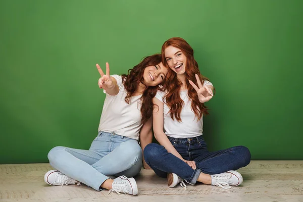 两个微笑的年轻红发女孩的肖像坐在地板上 显示和平姿态孤立的绿色背景 — 图库照片