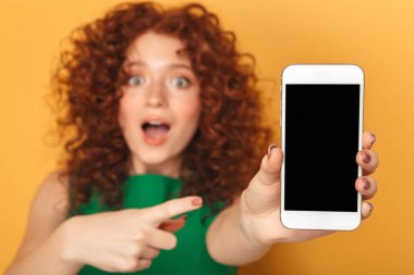 Elbise işaret parmağı, boş ekran cep telefonu üzerinde sarı arka plan izole bir sürpriz Kızıl saçlı kadın portresi