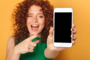 Elbise işaret parmağı, boş ekran cep telefonu üzerinde sarı arka plan izole bir mutlu kızıl saçlı kadın portresi
