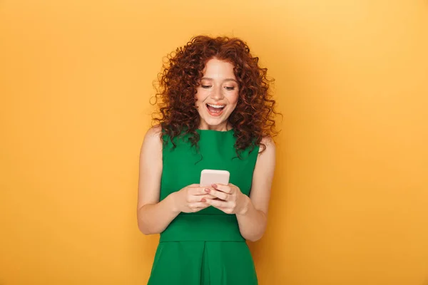 黄色背景に分離された携帯電話を使用してドレスを着た赤毛の陽気な女性の肖像画 — ストック写真