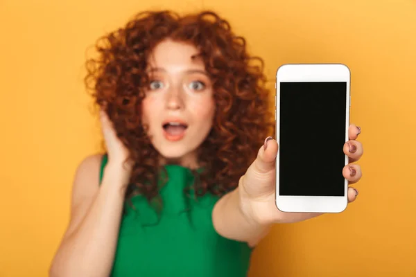黄色の背景に分離された空白の画面携帯電話を見せドレスでびっくりの赤毛の女性の肖像画 — ストック写真