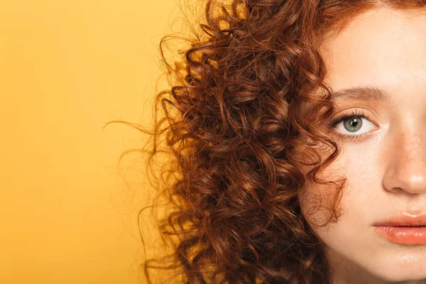 Half face close up portrait of a calm curly redhead woman looking at camera isolated over yellow background