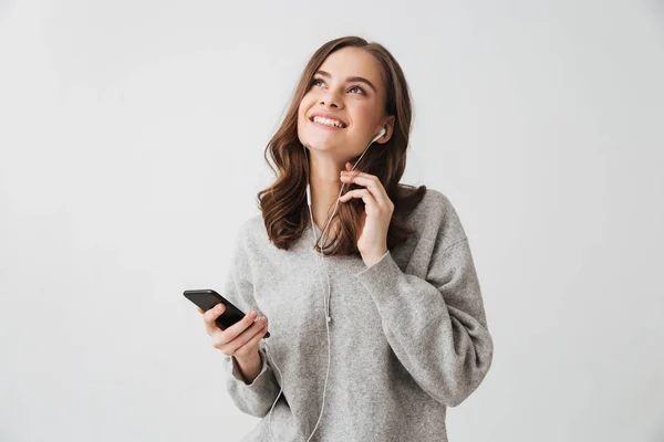 Pensive smiling brunette woman in sweater holding smartphone and looking up over grey background