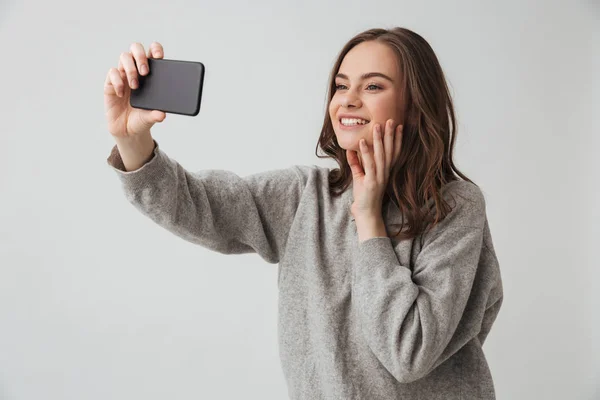 快乐的黑发妇女在毛衣做自拍在智能手机 而触摸她的脸灰色背景 — 图库照片
