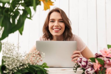 Genç Sevimli neşeli çiçekçi Bayan dizüstü bilgisayar kullanan Atölye çiçeklerle ayakta. Görünümlü kamera.