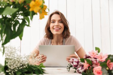Genç Sevimli neşeli çiçekçi Bayan dizüstü bilgisayar kullanan Atölye çiçeklerle ayakta. Görünümlü kamera.