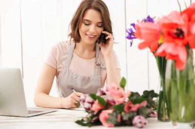 Genç Sevimli neşeli çiçekçi kadın çiçek notlar yazma cep telefonuyla konuşurken atölye üzerinde.