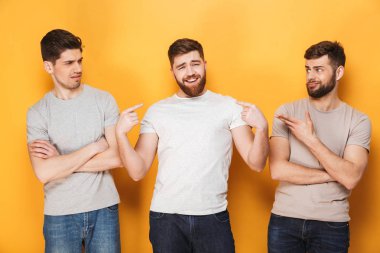 Two young confused men looking at their male friend isolated over yellow background clipart