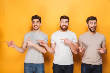 Üç genç erkekler uzak sarı arka plan üzerinde izole kopya alanı işaret heyecanlı