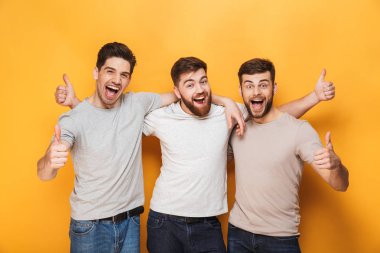 Üç genç heyecanlı adam başparmak sarı arka plan üzerinde izole gösterilmesini