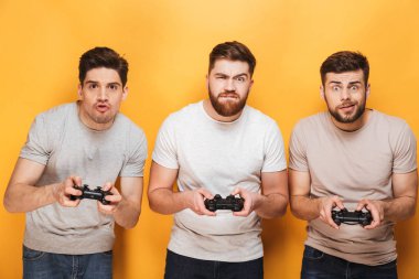 Üç genç heyecanlı adam sarı arka plan üzerinde izole video oyunları oynarken oyun çubukları holding