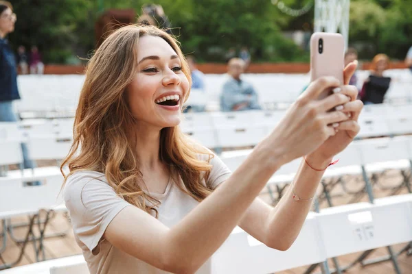 Smiling young girl taking picture with mobile phone while sitting on a chair at the park