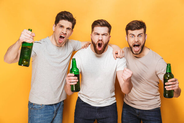 Three young joyful men holding beer and celebrating isolated over yellow background